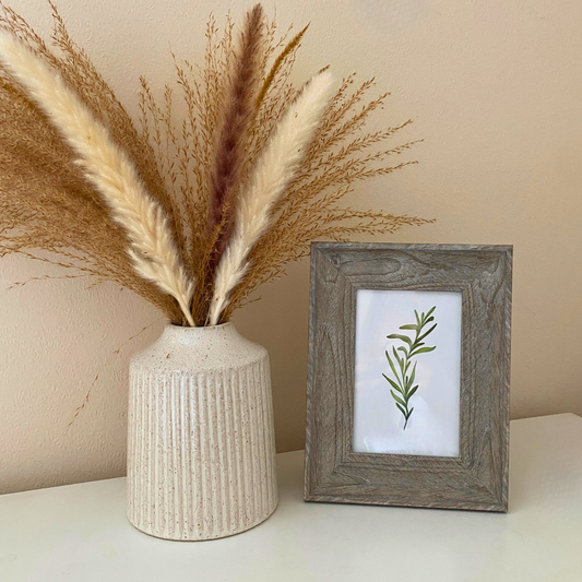 Decorative vase with dried pampas grass next to a framed print of a leaf on a neutral background
