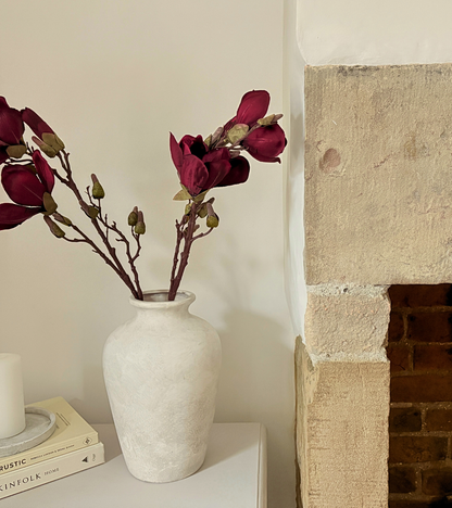 White vase with red flowers on a shelf next to a stone fireplace.